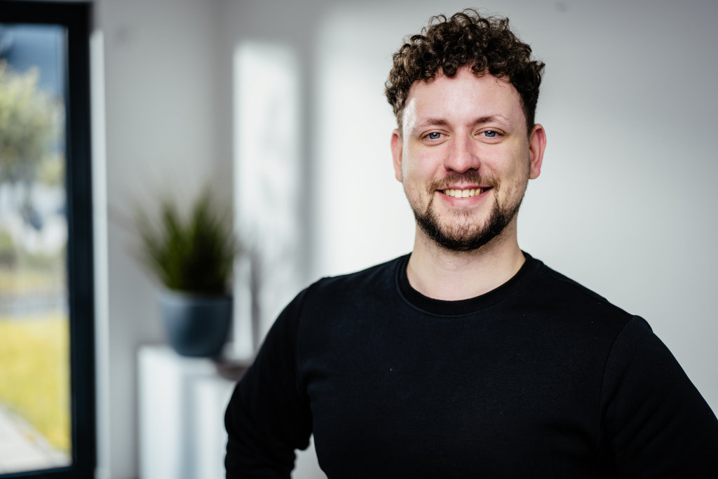 Portrait von jungem Mann mit braunem Haar und Bart und dunklem Oberteil.