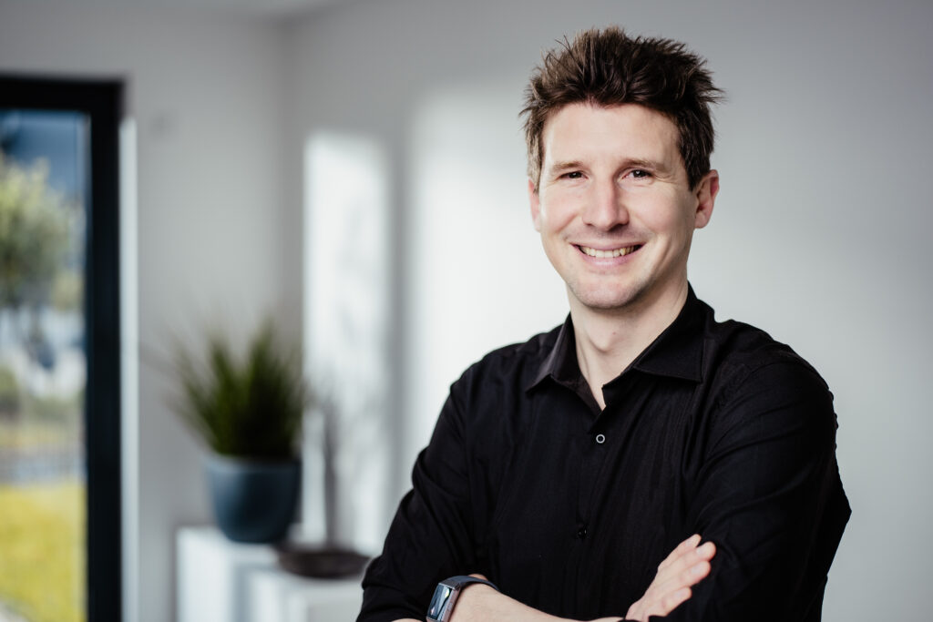 Portrait von jungem Mann mit braunem Haar und dunklem Oberteil der lächelnd die Arme verschränkt.