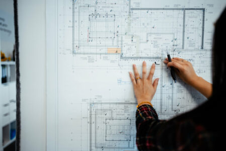 Frau steht vor Grundrissplan und hält Kugelschreiber in der rechten Hand.