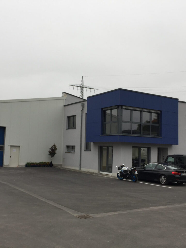 Weiße Lagerhalle vor der mehrere Fahrzeuge stehen.