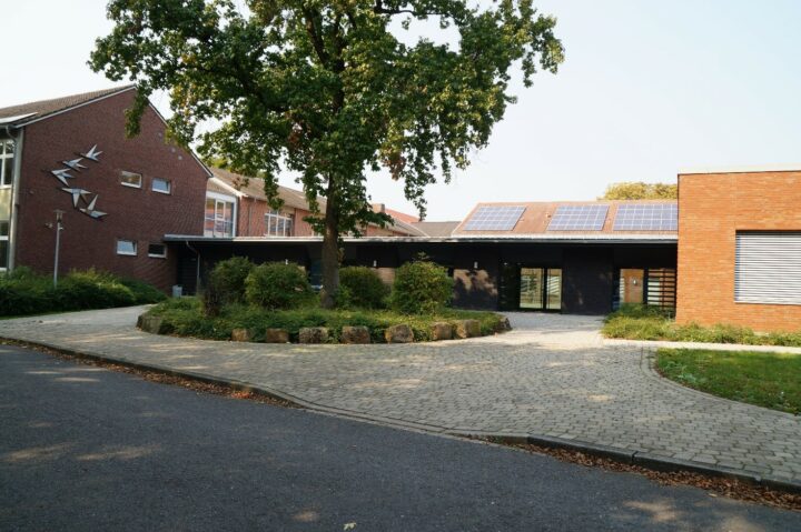 Eingangsbereich eines flachen Gebäudes vor dem ein rundes Beet mit grünem Baum steht.