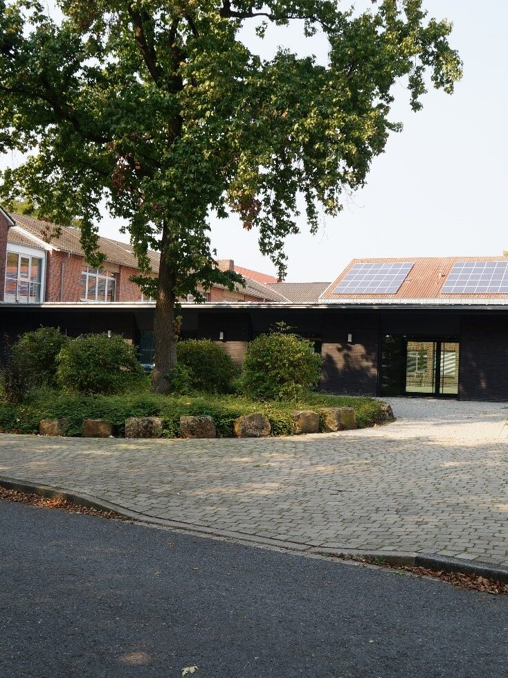 Eingangsbereich eines flachen Gebäudes vor dem ein rundes Beet mit grünem Baum steht.