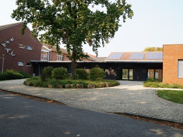 Eingangsbereich eines flachen Gebäudes vor dem ein rundes Beet mit grünem Baum steht.