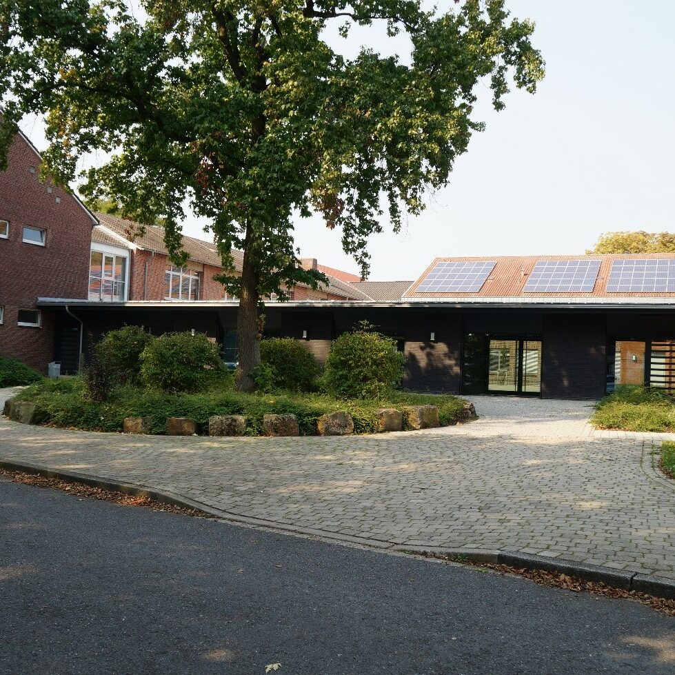 Eingangsbereich eines flachen Gebäudes vor dem ein rundes Beet mit grünem Baum steht.