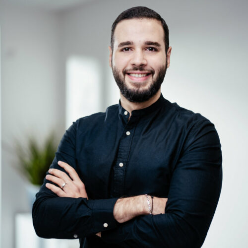 Mann mit kurzen braunen Haaren und vollem Bart in schwarzem Hemd verschränkt die Arme und lächelt freundlich