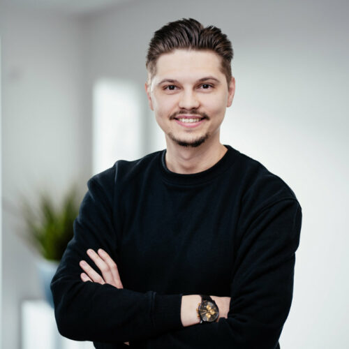 Junger Mann mit hochgegelten braunen Haaren, Oberlippenbart und dunklem Pullover lächelt selbstbewusst