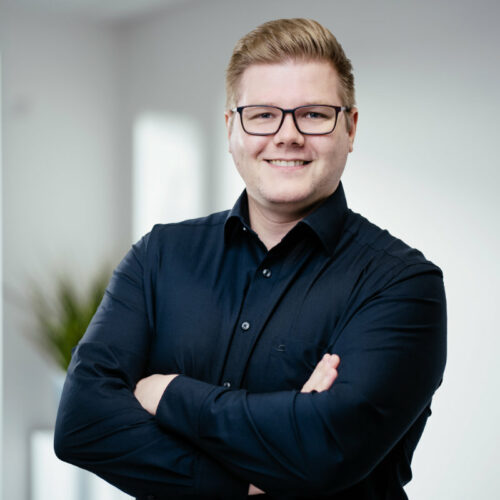 Mann mit blonden Haaren, Brille und dunklem Hemd lächelt freundlich