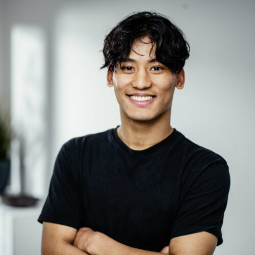 Dünner, junger Mann mit längeren schwarzen Haaren und schwarzem T-Shirt lächelt freundlich