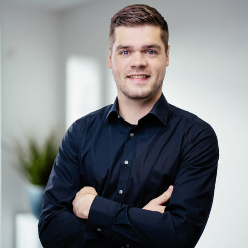 Großer Mann mit dunkel-blonden Haaren, dunkelblauem Hemd verschränkt die Arme und lächelt freundlich