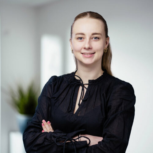 Junge Frau mit langen dunkel-blonden Haaren, goldenen Ohrringen und dunkler Bluse lächelt freundlich