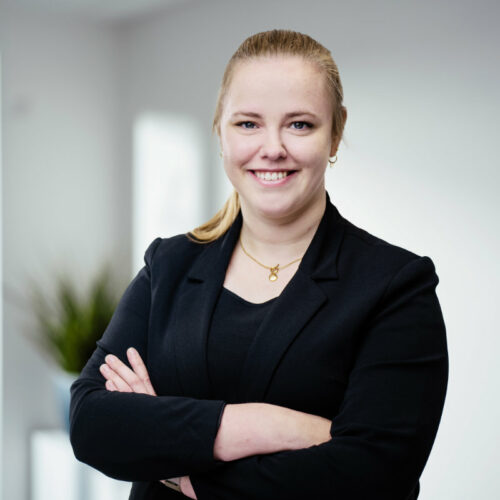 Junge Frau mit blondem Pferdeschwanz, goldener Halskette und dunklem Blazer lächelt freundlich