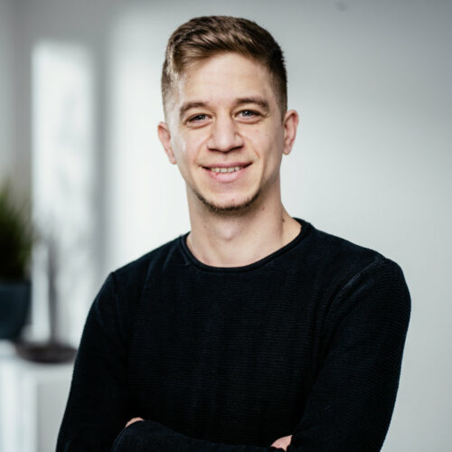 Dünner junger Mann mit dunkel-blonden Haaren und dunklem Pullover lächelt freundlich