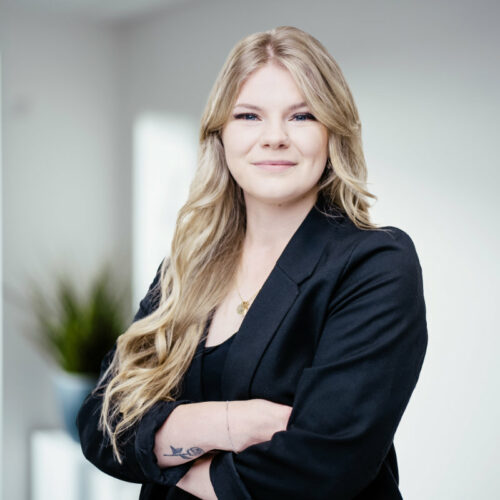 Junge Frau mit offenen, langen blonden Haaren und dunklem Blazer lächelt freundlich