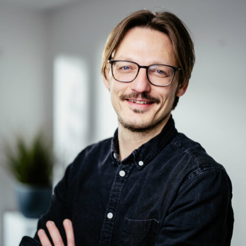 Mann mit braunem Mittelscheitel, Brille und dunklem Jeanshemd lächelt freundlich