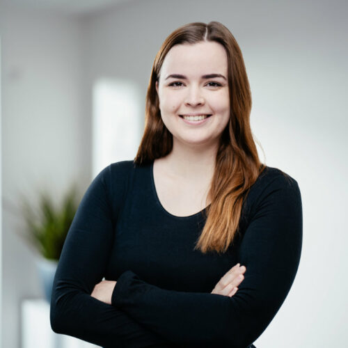 Junge Frau mit offenen, langen dunkel-blonden Haaren verschränkt die Arme und lächelt freundlich