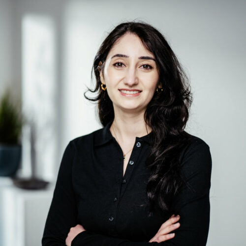 Dünne junge Frau mit goldenen Ohrringen und offenen, dunklen Haaren verschränkt die Arme und lächelt freundlich