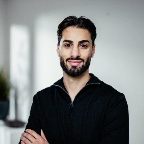 Dünner junger Mann mit schwarzen Haaren, Vollbart und dunklem Reisverschlusspullover lächelt