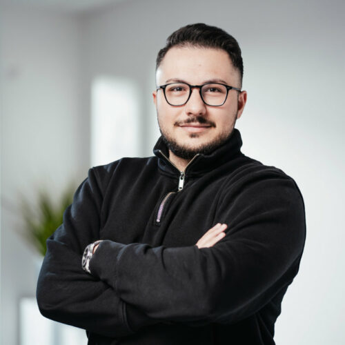 Mann in Brille mit brünettem Haar und Bart verschränkt die Arme und lächelt